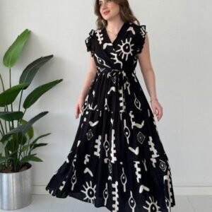 Woman wearing a black printed maxi dress with white abstract patterns and ruffle sleeves, posing indoors near a plant.