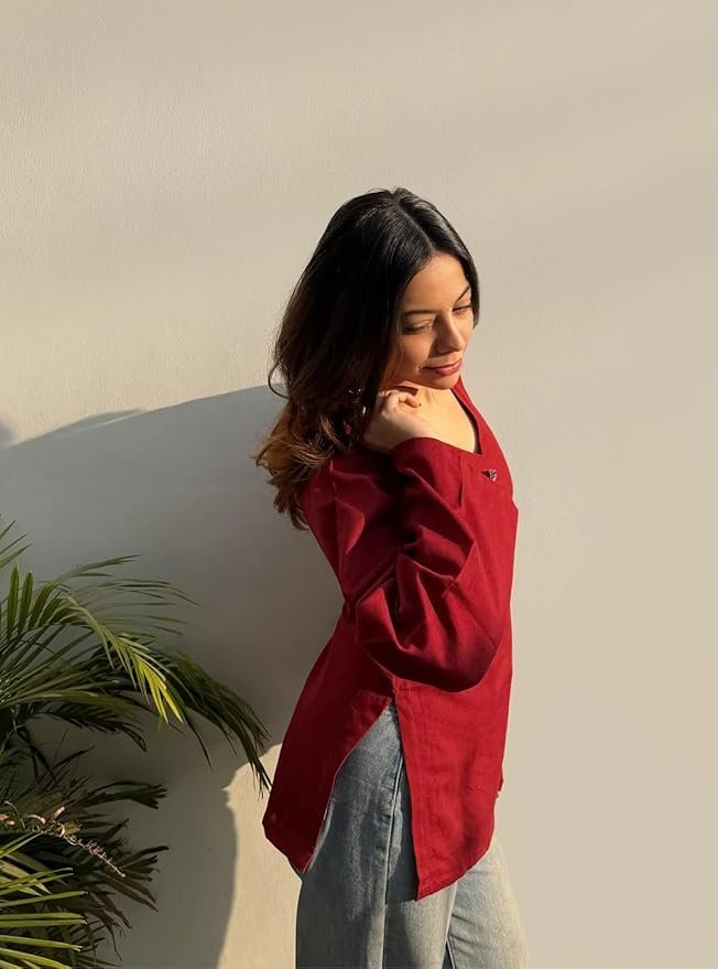 Woman wearing a stylish maroon V-neck top with flared sleeves and grey pants, posing in natural sunlight against a minimal white wall.
