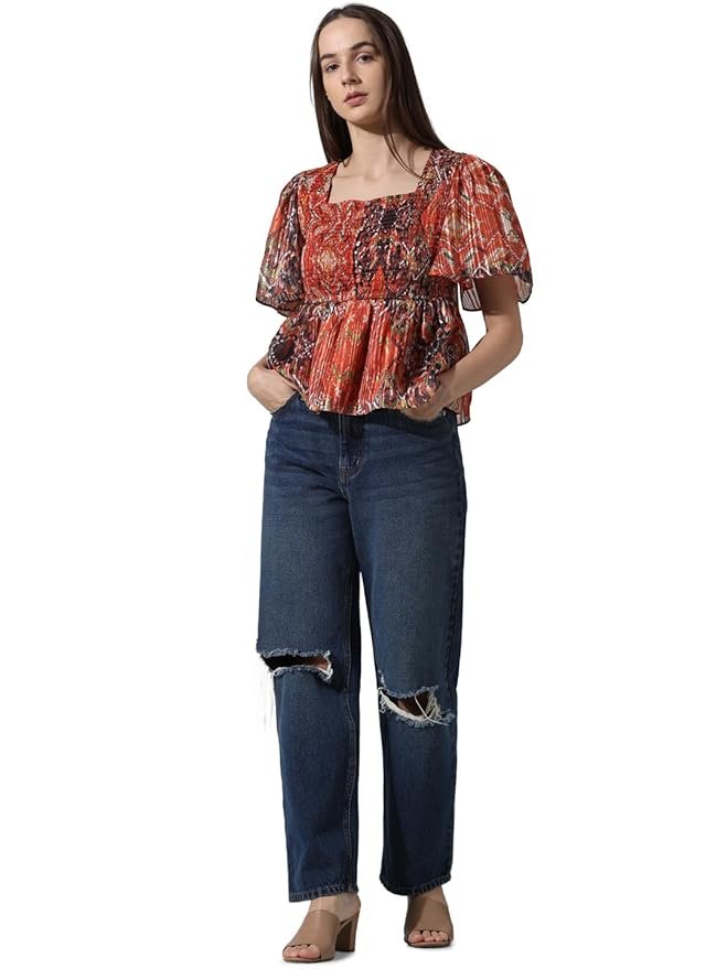 Woman wearing a red printed puff sleeve peplum top paired with distressed blue jeans and heels against a plain background.