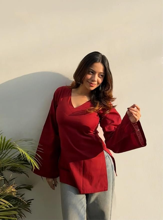 Woman wearing a stylish maroon V-neck top with flared sleeves and grey pants, posing in natural sunlight against a minimal white wall.