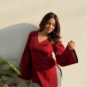 Woman wearing a stylish maroon V-neck top with flared sleeves and grey pants, posing in natural sunlight against a minimal white wall.